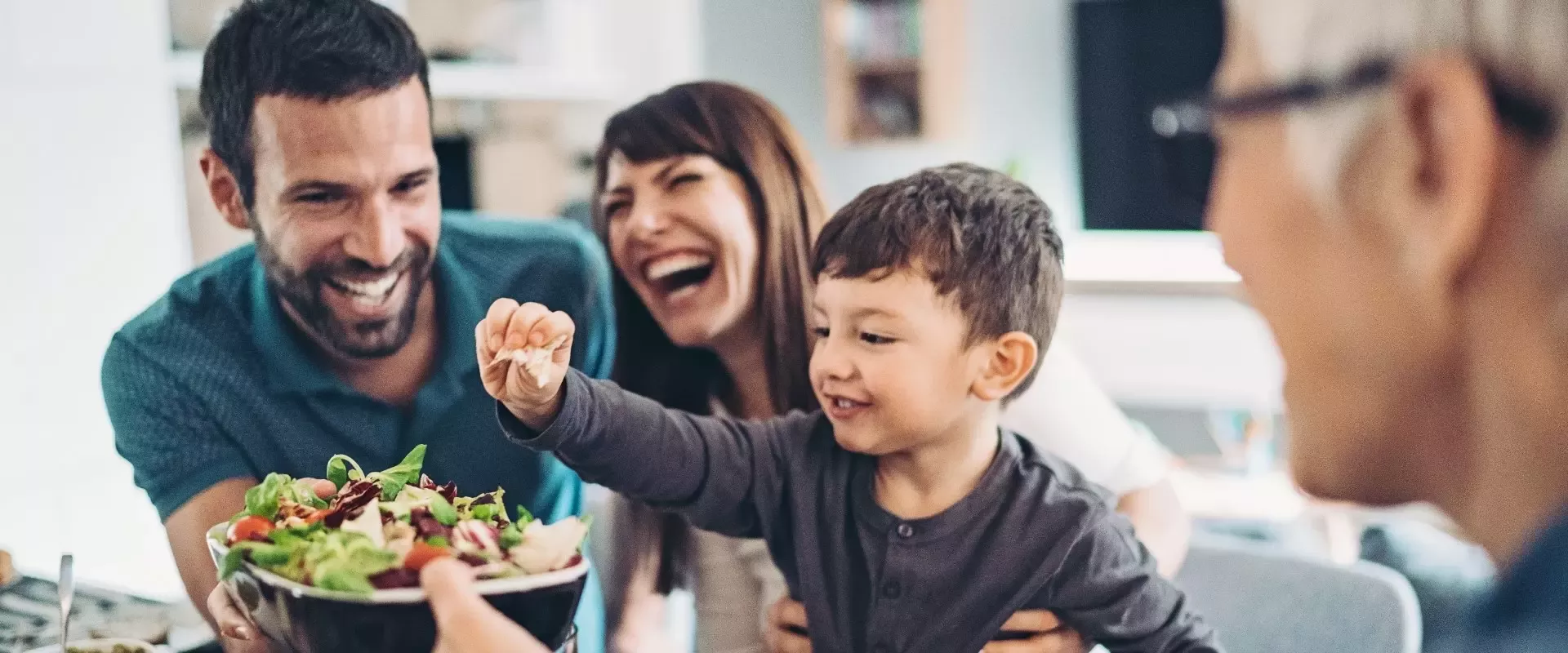 Pai, mãe e filho em redor da mesa a sorrir enquanto a avó segura a taça de salada